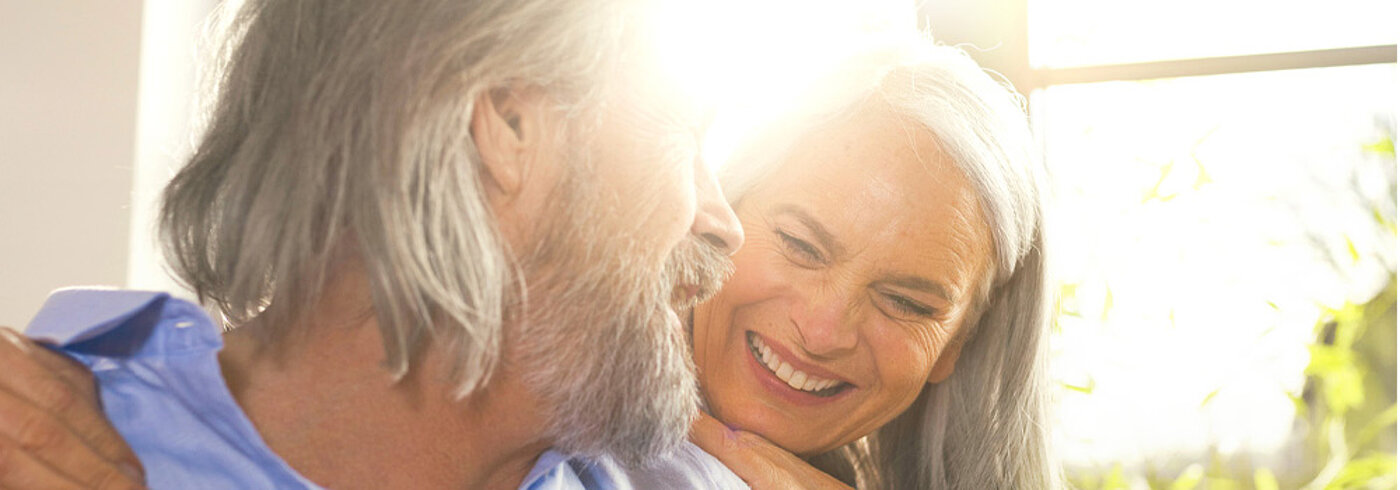 Ein Mann und eine Frau mit grauen Haaaren lächeln sich verliebt an. Im Hintergrund erstrahlt die Sonne.